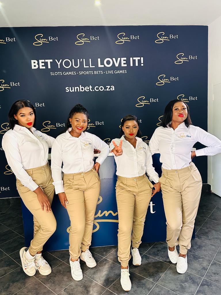 Isilumko-Activate Four instore brand ambassadors pose in front of a sun bet branded backdrop, wearing matching beige uniforms and red lipstick.