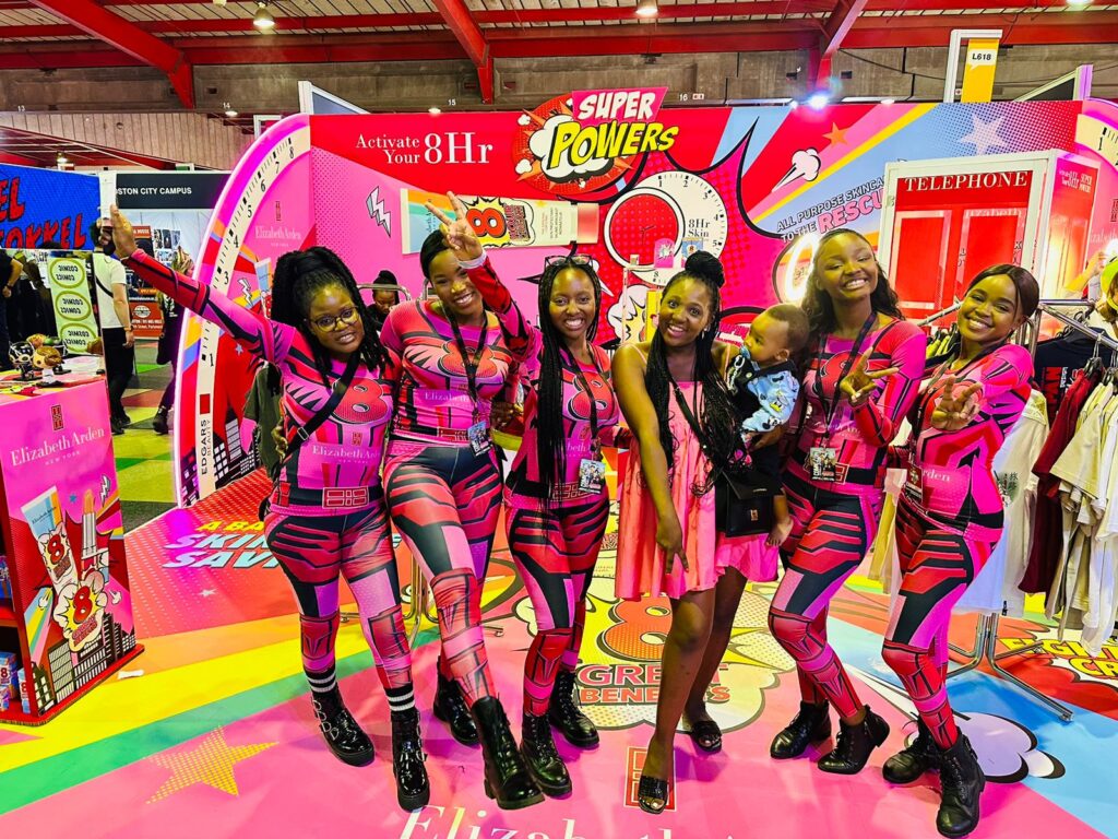 Isilumko-Activate Six instore brand ambassadors in matching pink and black superhero costumes, smiling and posing with raised arms at a colorful exhibition booth.